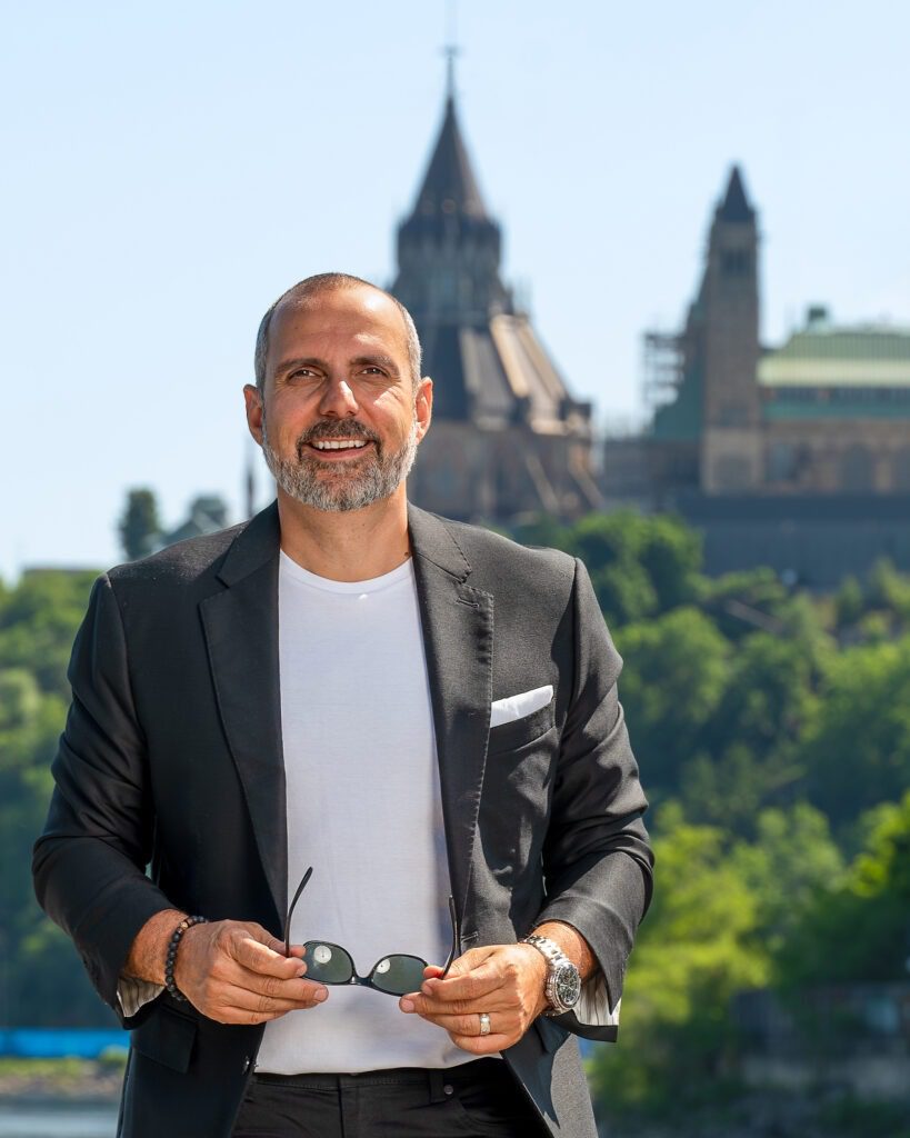 Environmental Headshot of John Zinati, sunglasses in hand, with the Parliament of Canada building in the background