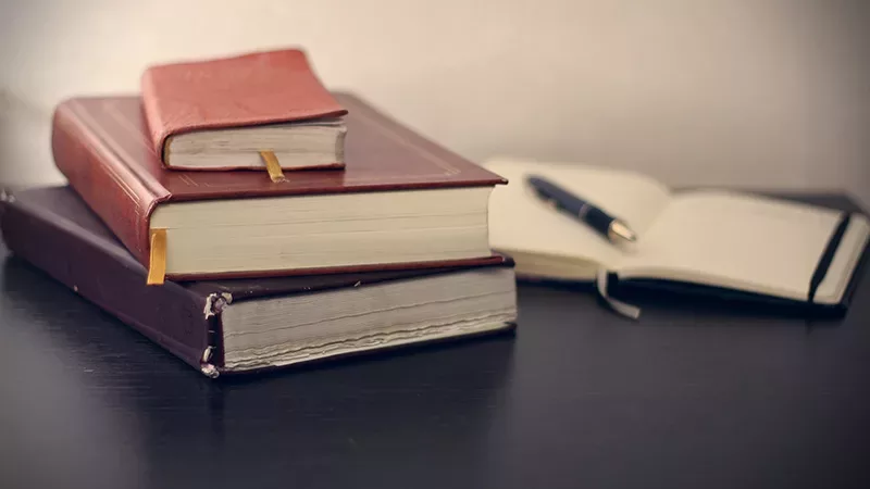 Stock image of three books stacked with an open notebook in the background that has a pen placed over it.